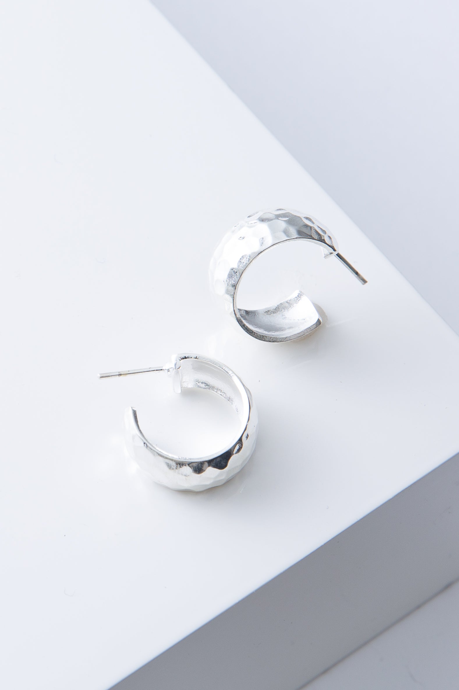 These are chunky, demi-hoop earrings that are open at the back. They are composed of flattened silver plated bronze with a hammered texture. The hoops are attached to a steel post.
