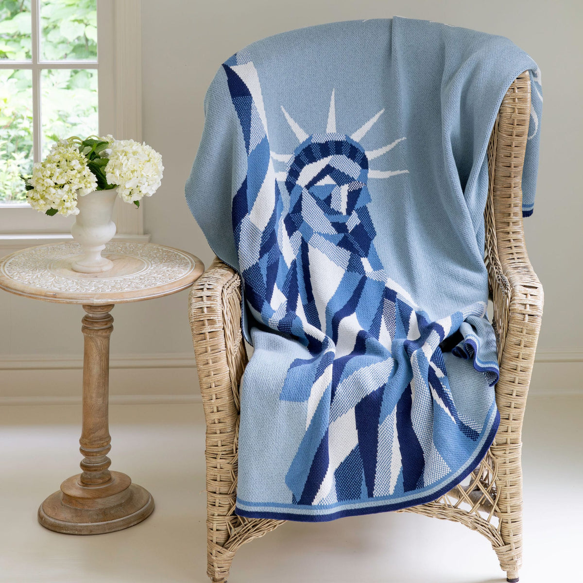 The in2green Statue of Liberty | We the People Cotton Sustainable Knit Modern Throw Blanket, featuring an eye-catching blue and white design that honors American identity, is artfully draped over a wicker chair beside a small table topped with a vase of white flowers by the window.