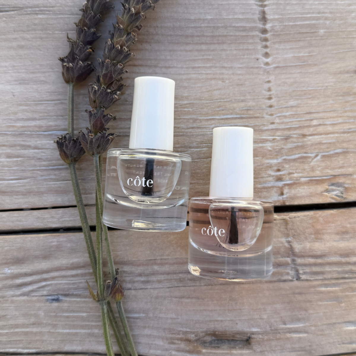Two clear nail polish bottles with white caps on a wooden surface next to lavender.