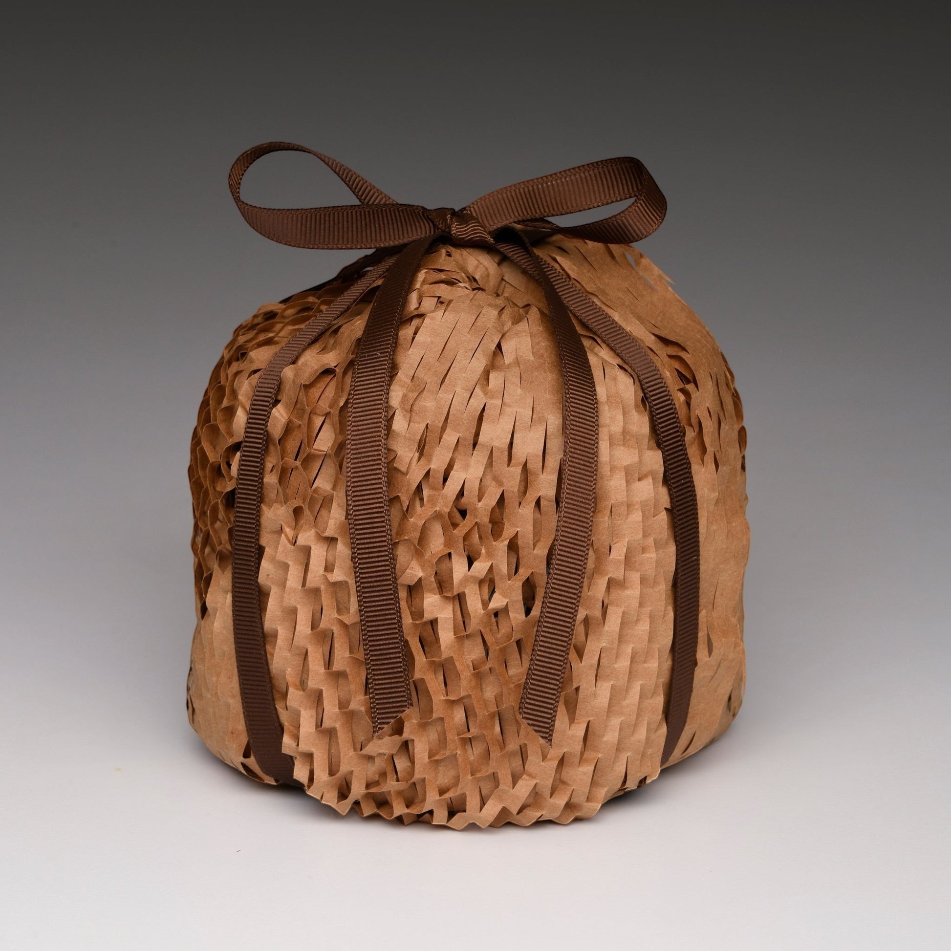 A round, hand-woven basket or pouch made of light brown natural fibers, tied at the top with a dark brown ribbon bow, set against a neutral gray gradient background.