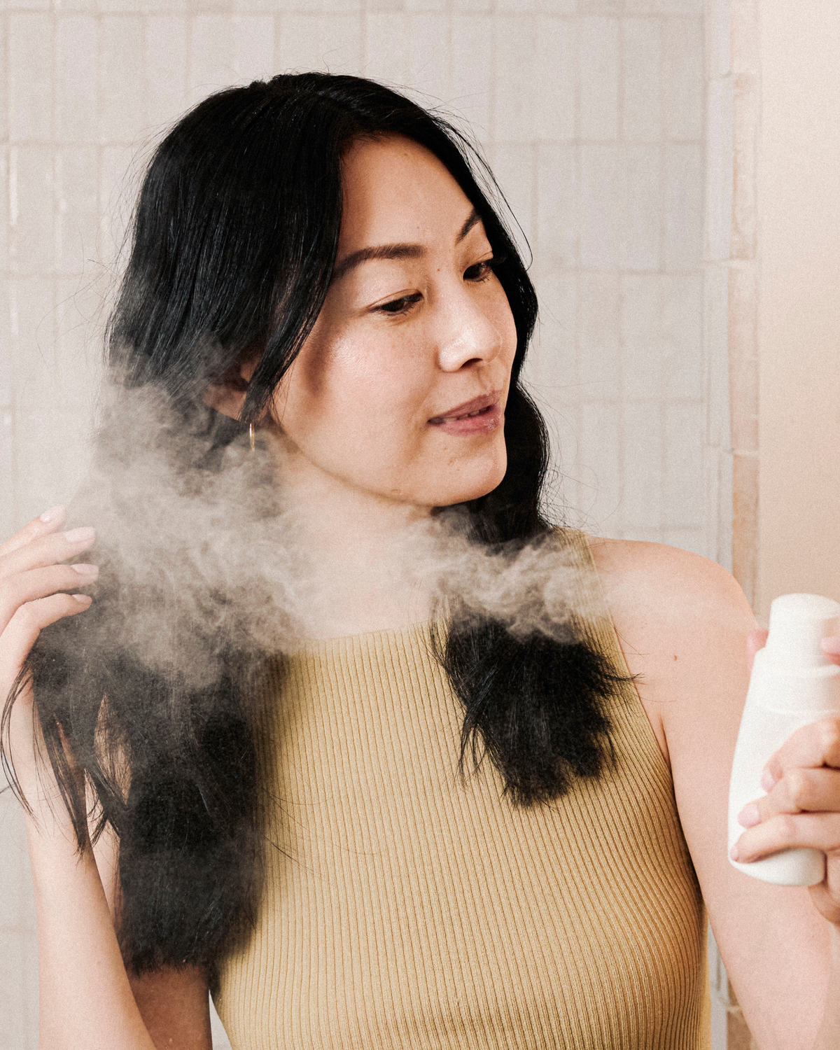 Woman using a hair product with steam coming from her hair, against a neutral background.