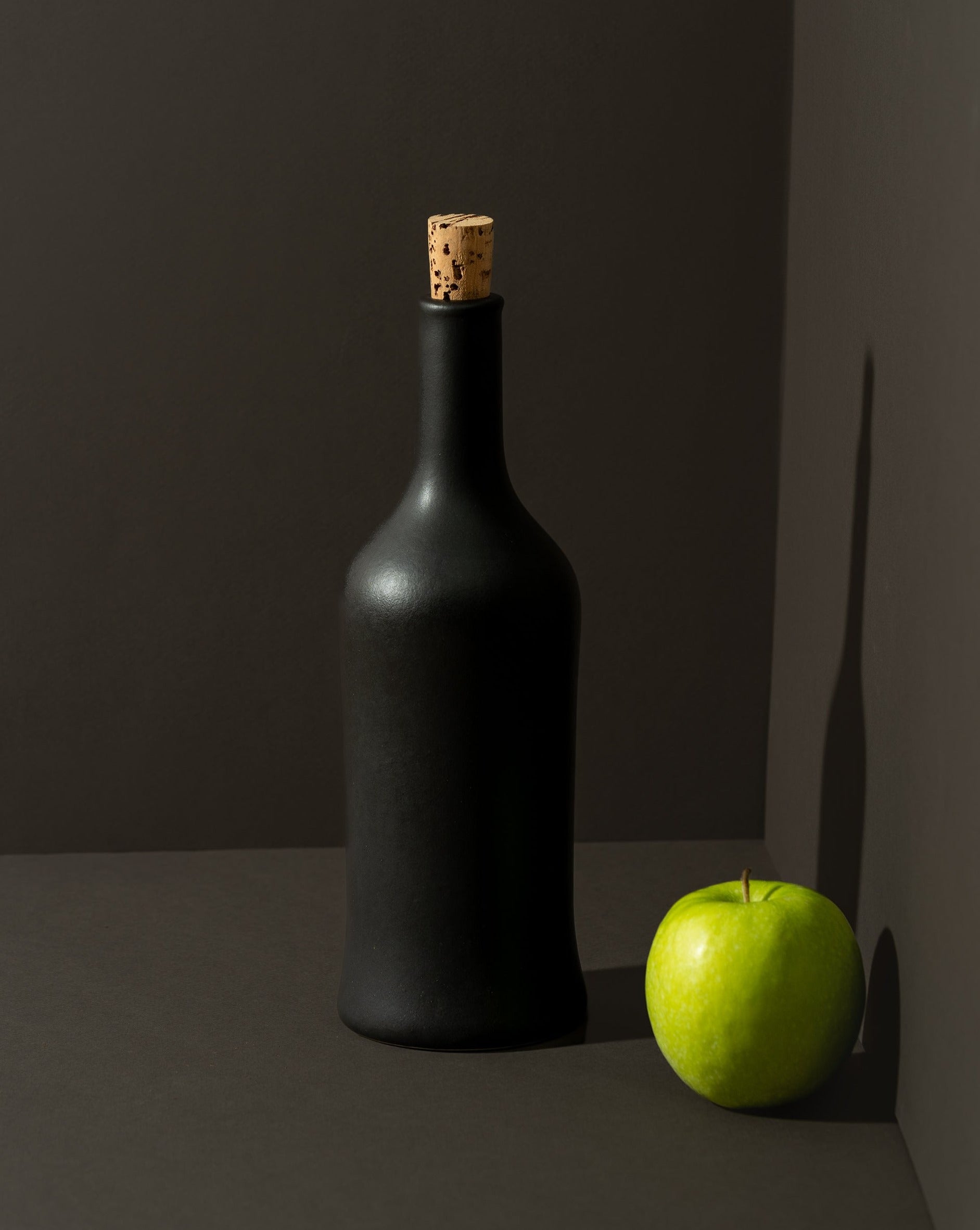 A white ceramic olive oil dispenser with a cork stopper, displayed next to a garlic bulb, on a neutral background.