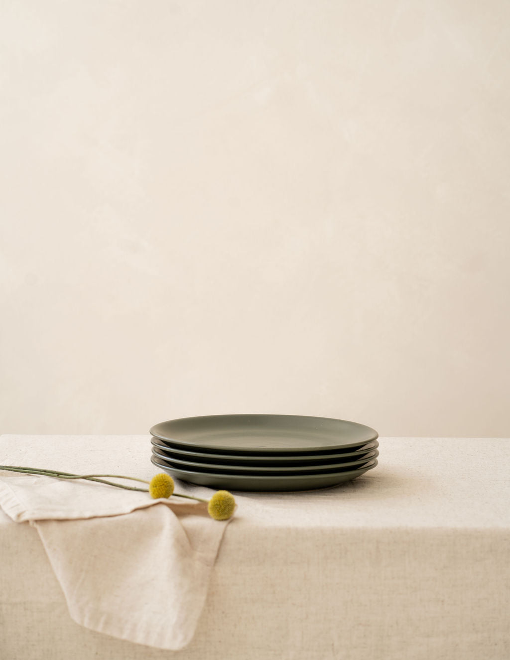 Moss green non-toxic ceramic dinner plates from Hamkke Home stacked on a neutral linen tablecloth with yellow billy button stems.