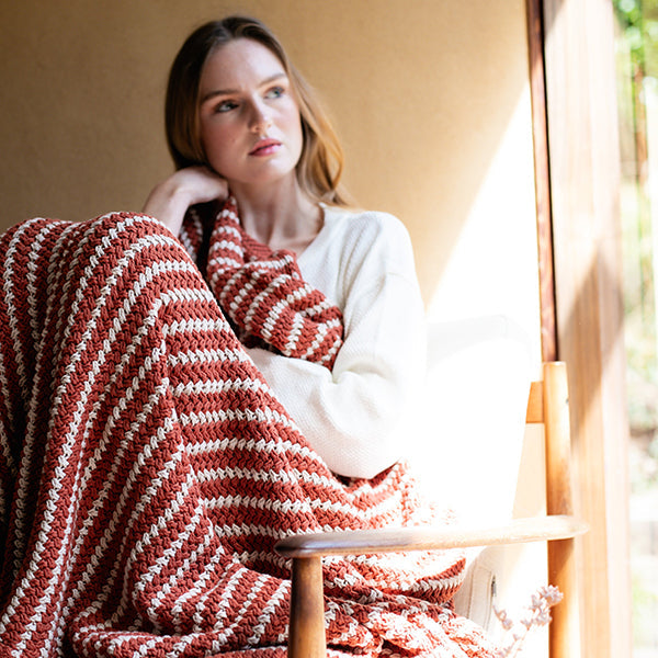 Woman sitting on a wooden chair with a red and white striped cotton throw blanket draped over her, in a softly lit room.