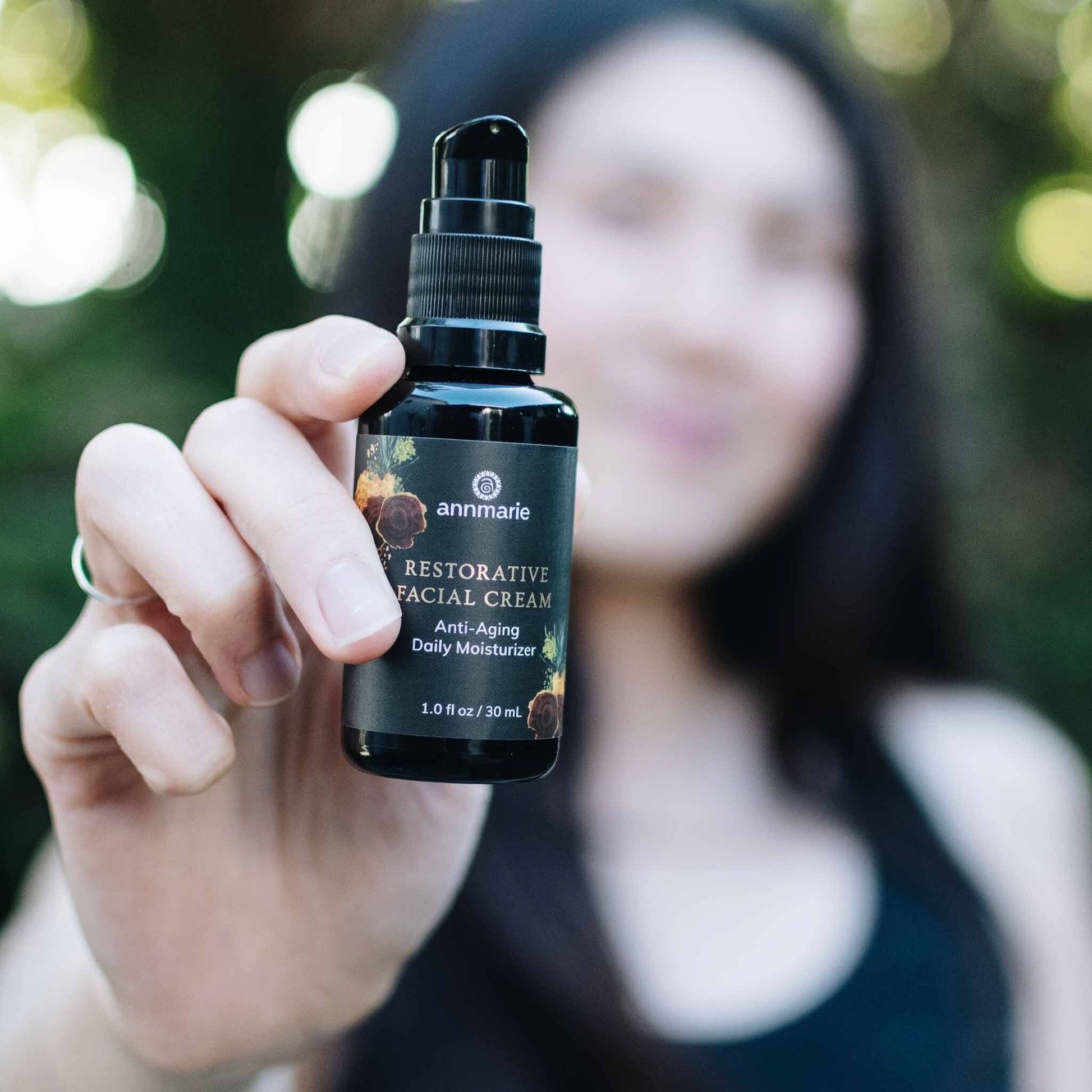 Person holding Annmarie Restorative Facial Cream anti-aging daily moisturizer bottle outdoors with soft greenery in the background.