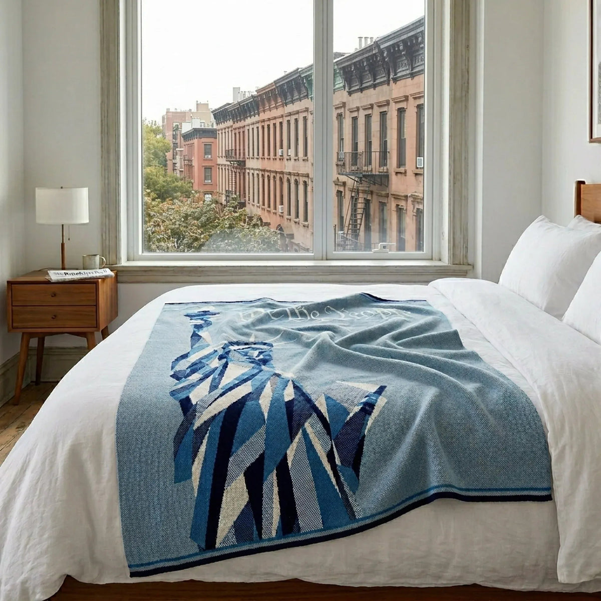 A neatly made bed featuring the in2green Statue of Liberty | We the People cotton sustainable knit modern throw blanket and a patriotic accent is positioned in front of a large window with views of city brownstone buildings.