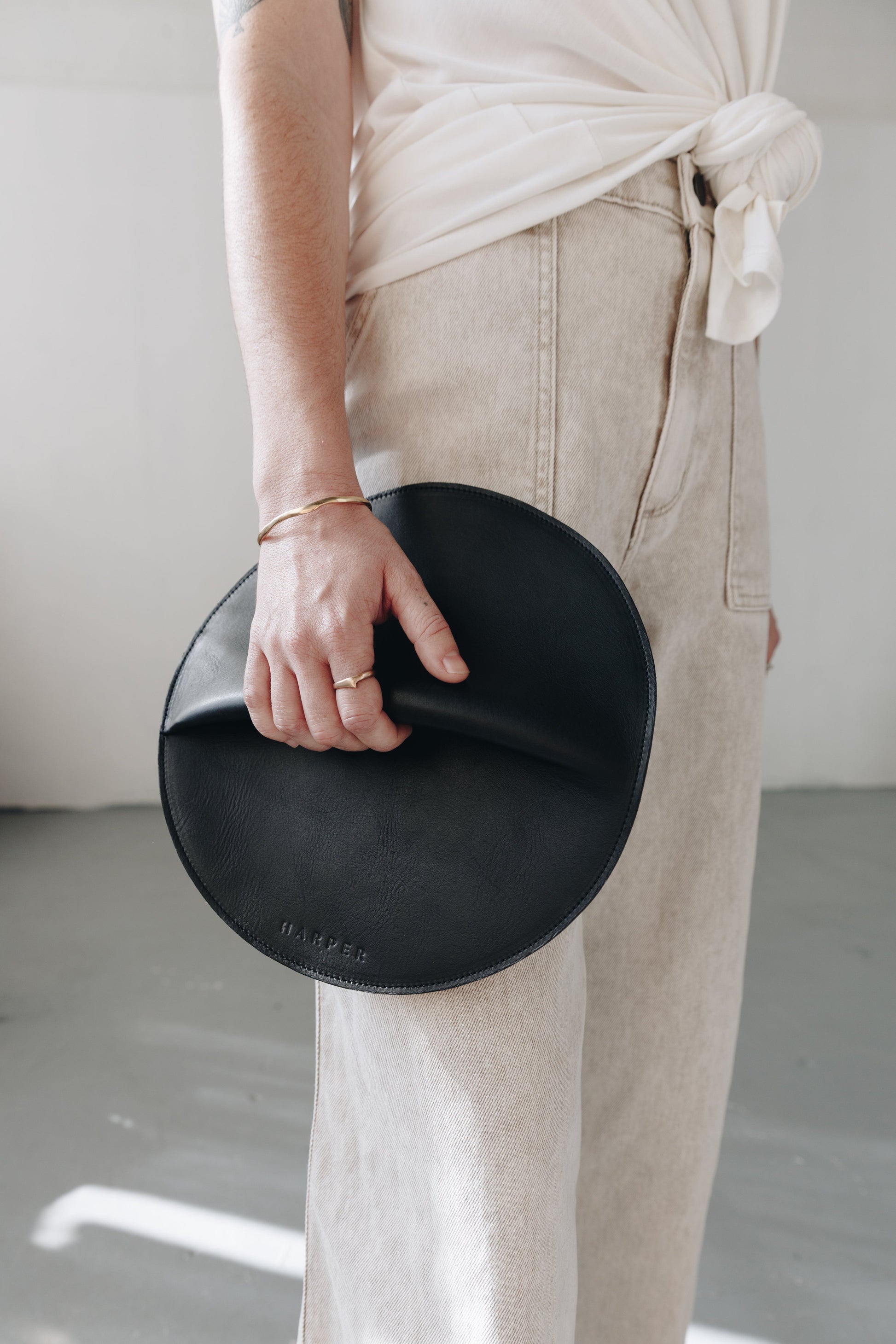 A woman's arms are visible, holding a circular black leather clutch with the reverse side and zipper visible. The zipper includes a matching zipper pull.
