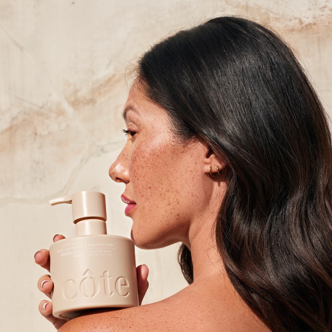 Woman with bottle of Côte Beauty Nourishing Daily Shampoo Non-Toxic Sulfate-Free Hair Cleanser on her shoulder. 