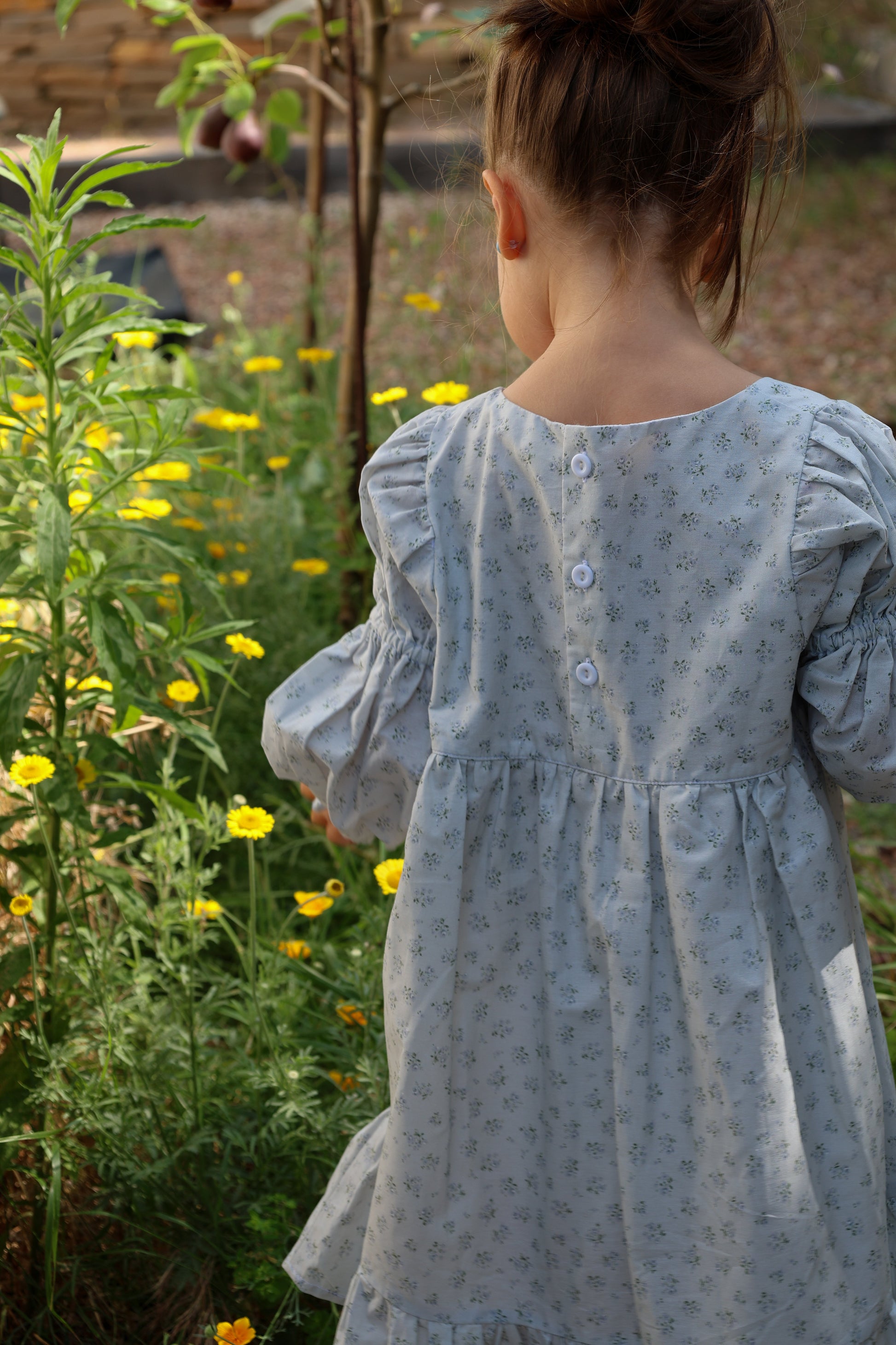 Child in a light blue dress standing in a garden with yellow flowers.