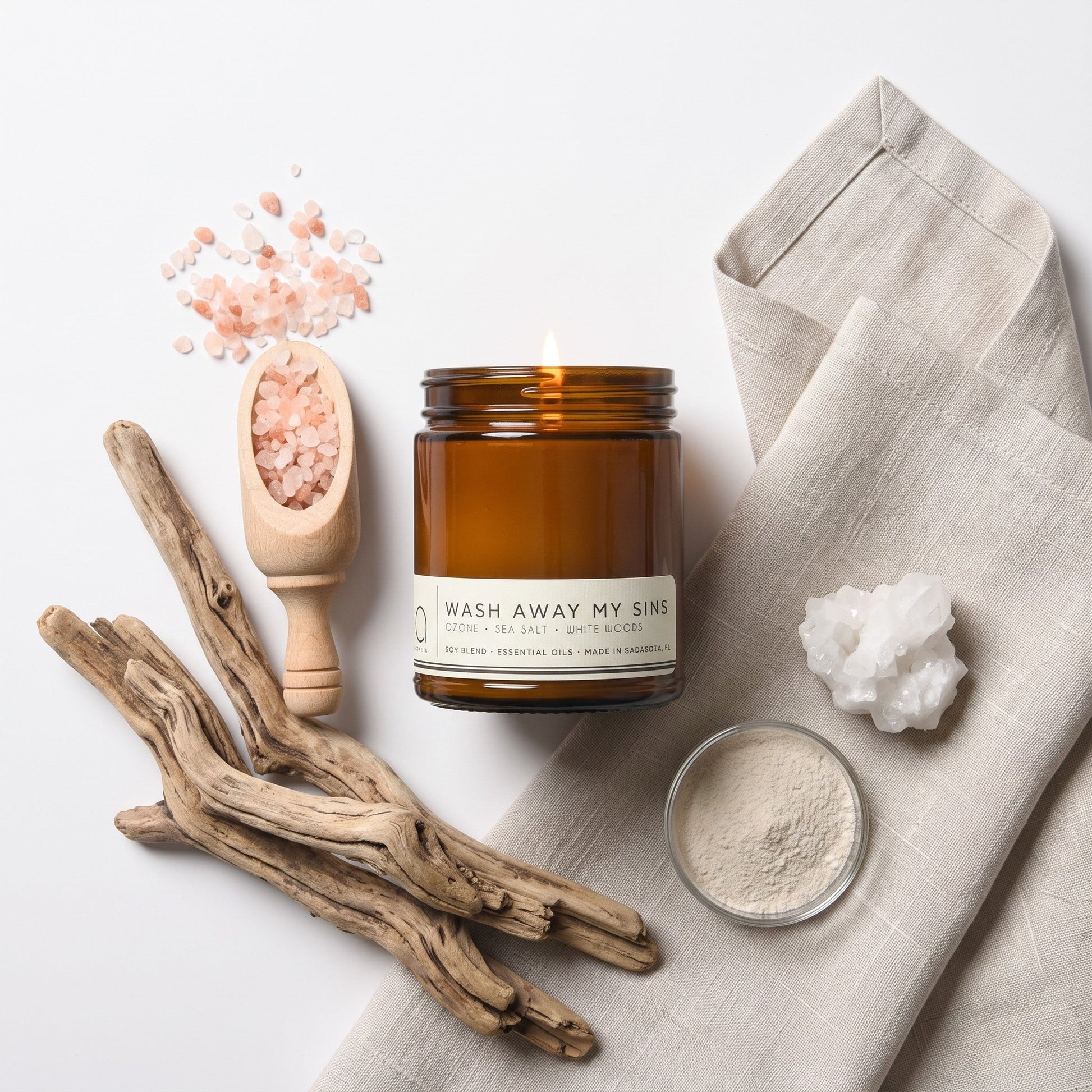 An amber glass jar candle with a white label reading 'WASH AWAY MY SINS' is styled in a flat lay with pink salt crystals, a wooden scoop, driftwood, a folded linen cloth, and a small dish of white powder on a white background.