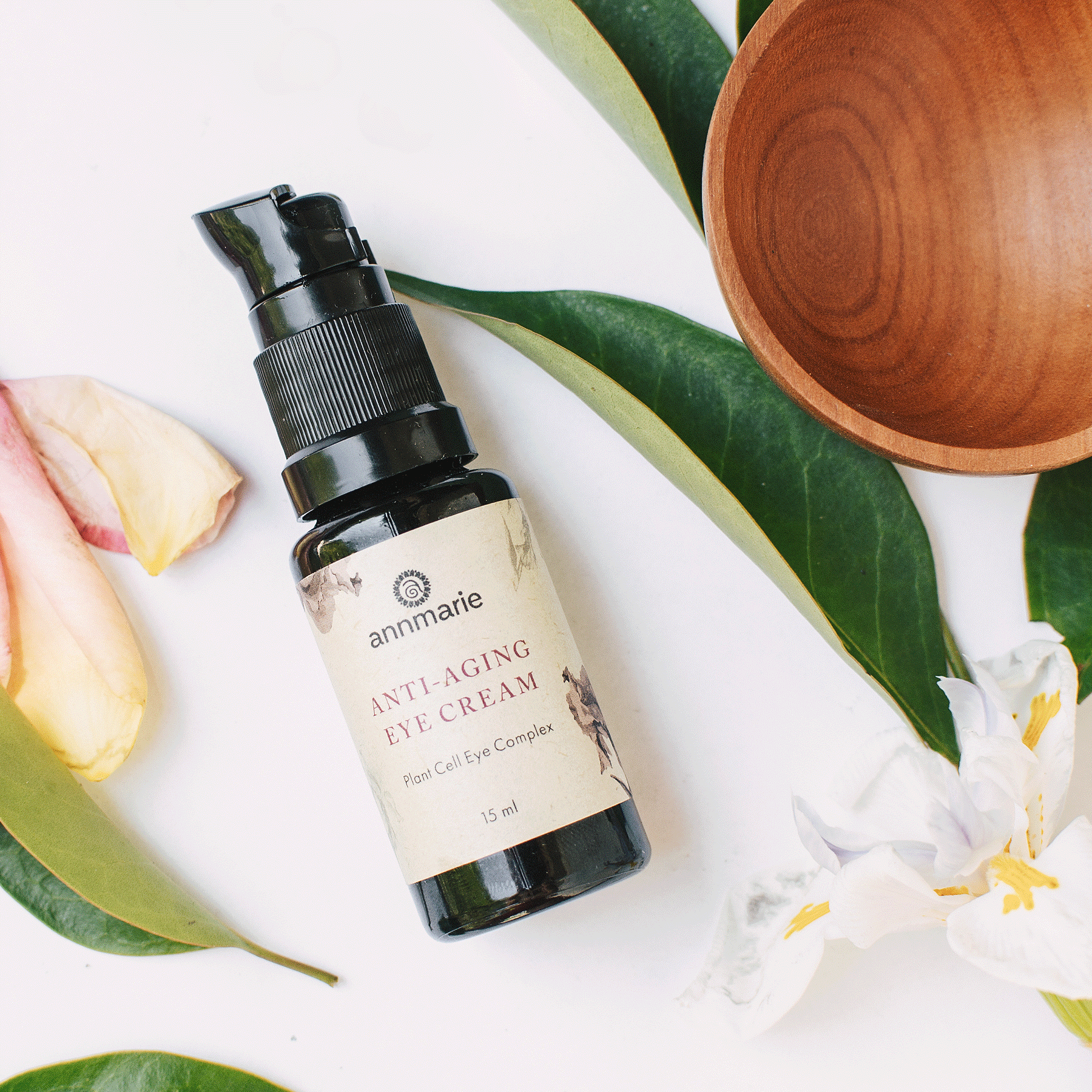 Anti-Aging Eye Cream bottle styled on a white surface with green leaves, soft flower petals, and a wooden bowl for a natural skincare aesthetic.