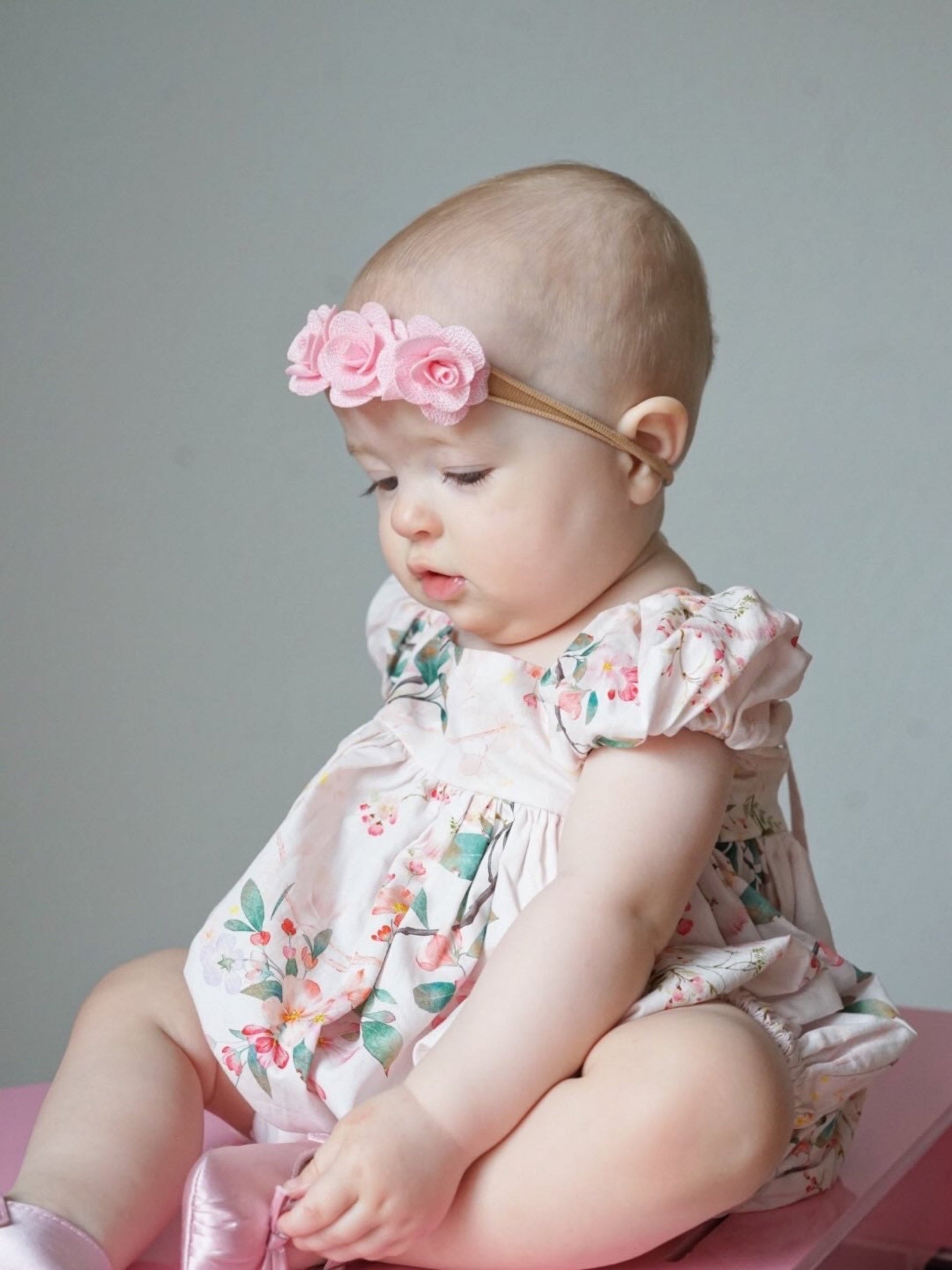 Baby wearing a floral Bubble and headband sitting on a pink surface with a gray background