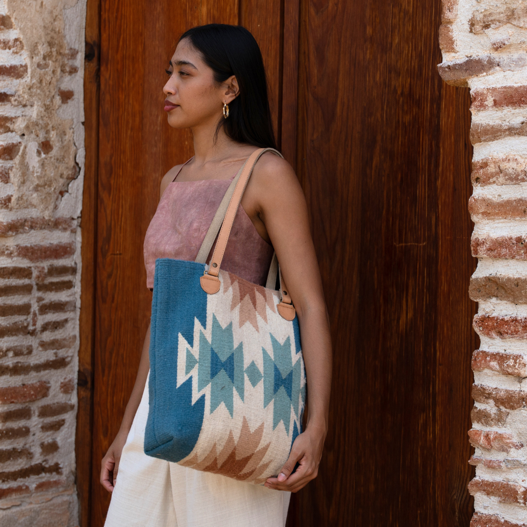 Woman carrying handwoven bucket bag in rose and blue with abstract diamond and lightning pattern, leather shoulder straps, and relaxed shape, ideal for everyday or beach use.