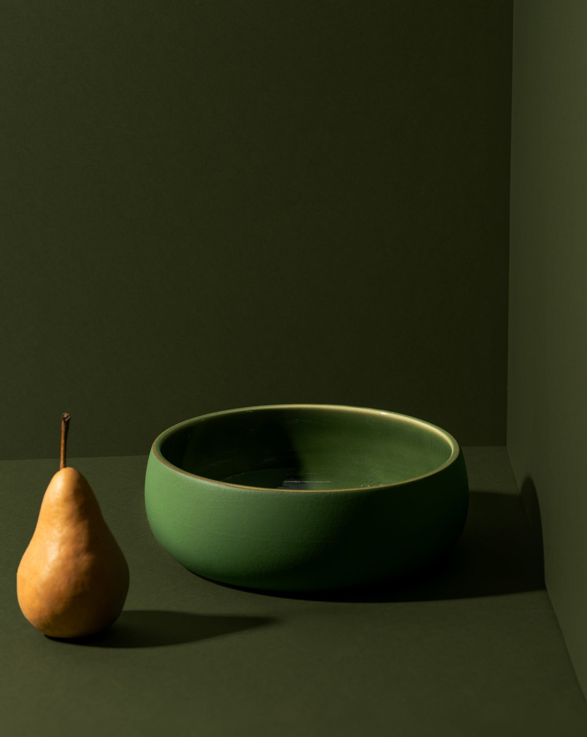 A matte green stoneware serving bowl displayed against a matching green background, with an oval-shaped fruit standing upright beside it, adding a natural touch to the composition.

