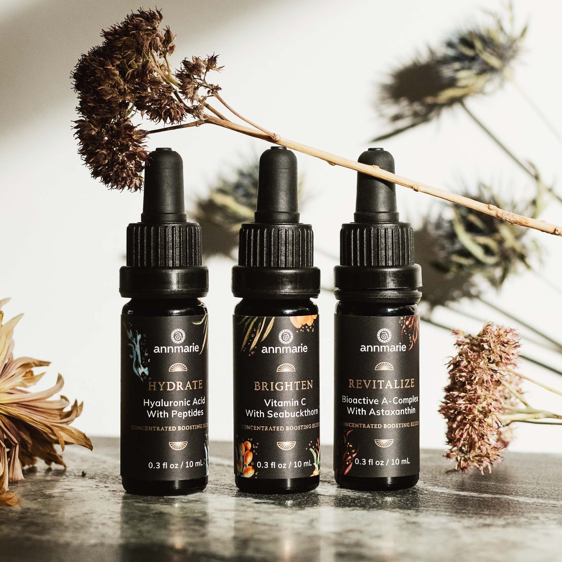 Three concentrated boosting elixirs—Hydrate, Brighten, and Revitalize—displayed in dropper bottles with dried botanical stems around them.