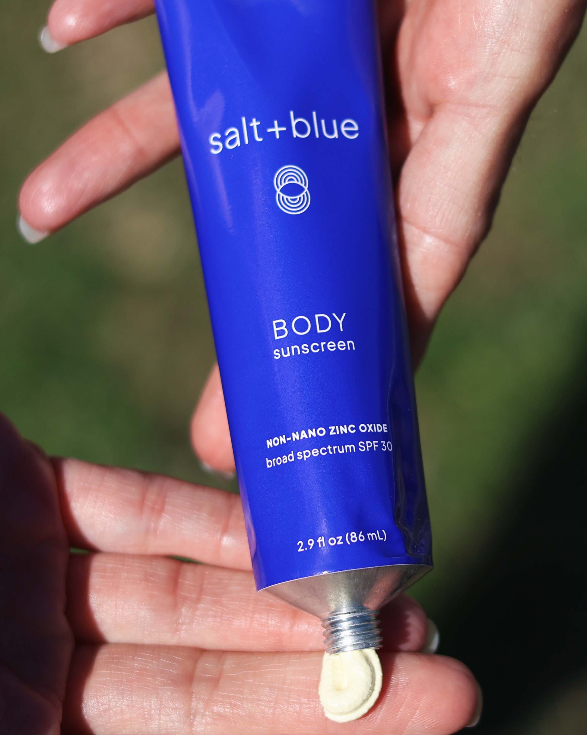 Blue tube of salt+blue body sunscreen held in a hand with a blurred green background