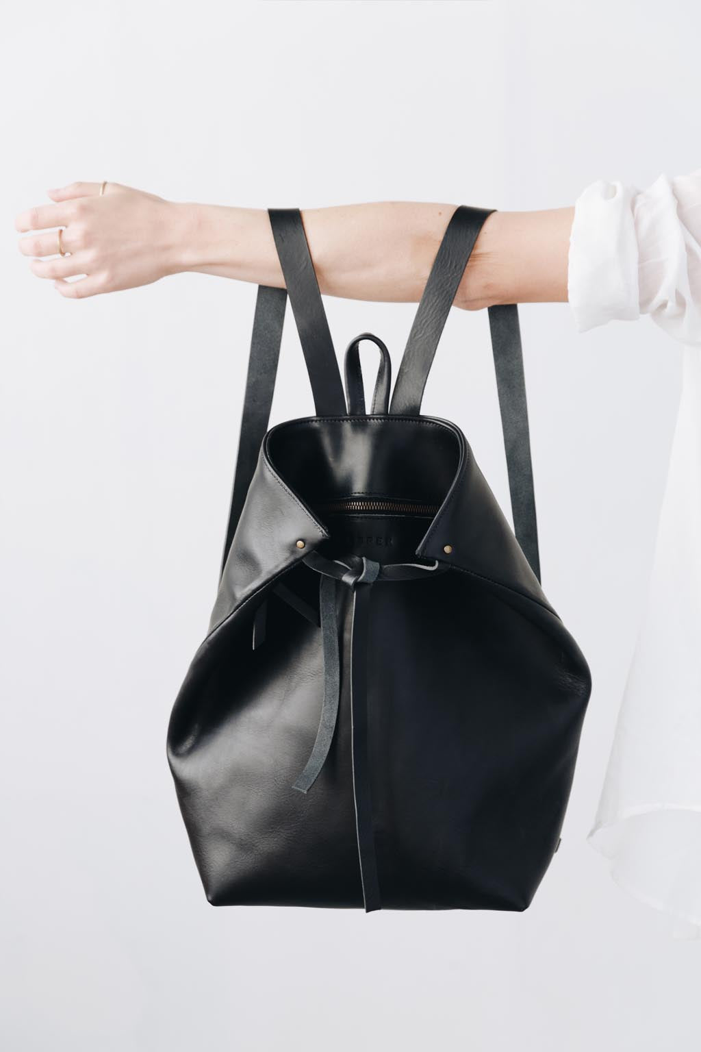 A woman faces away from the camera, wearing a black leather backpack. The top corners of the backpack are cinched with straps that knot over a zipper, creating a unique shape.