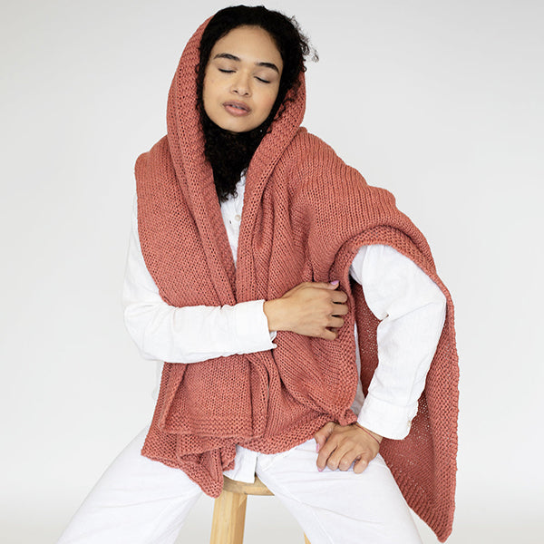 Woman wearing a rosewood pink knitted cotton throw around her head over a white shirt on a white background.