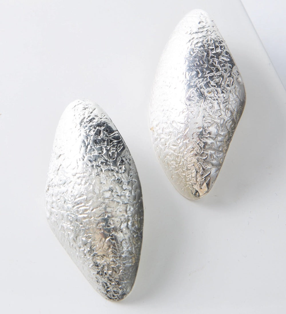These are post-style earrings composed of shining silver-plated brass with a hammered texture. The earrings have an irregular, organic look and are roughly diamond shaped, with smooth rounded edges.