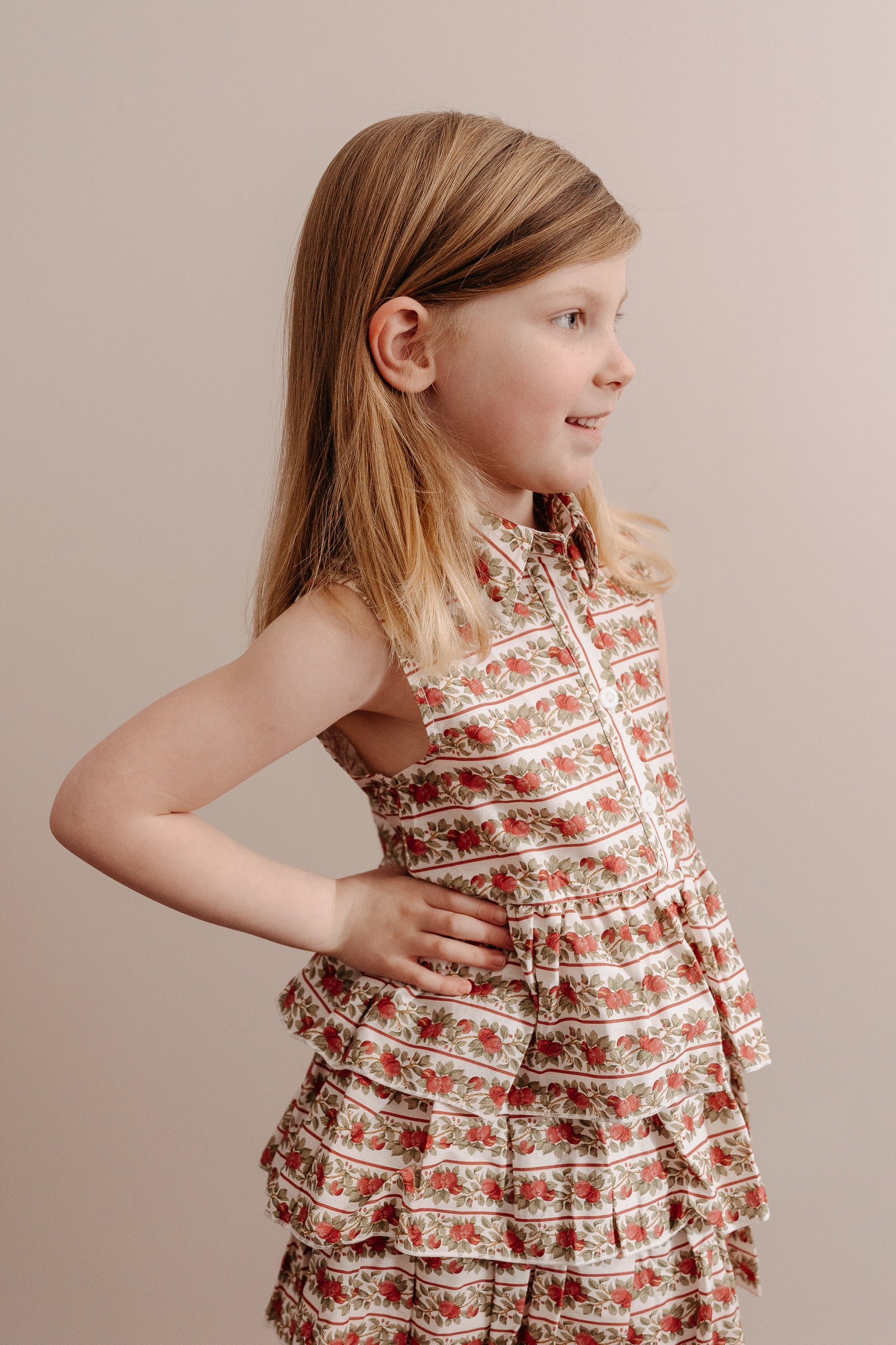 A young girl wearing a sleeveless, floral print, button-down, multi-tiered ruffle skirt dress.