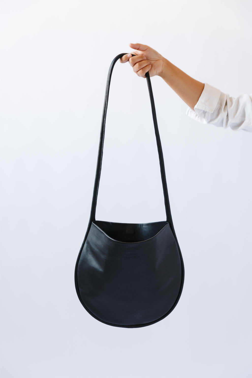 A woman's arm is visible, holding out a black leather shoulder bag by the thin strap. The leather is soft and shiny, and the bag is a half moon shape.