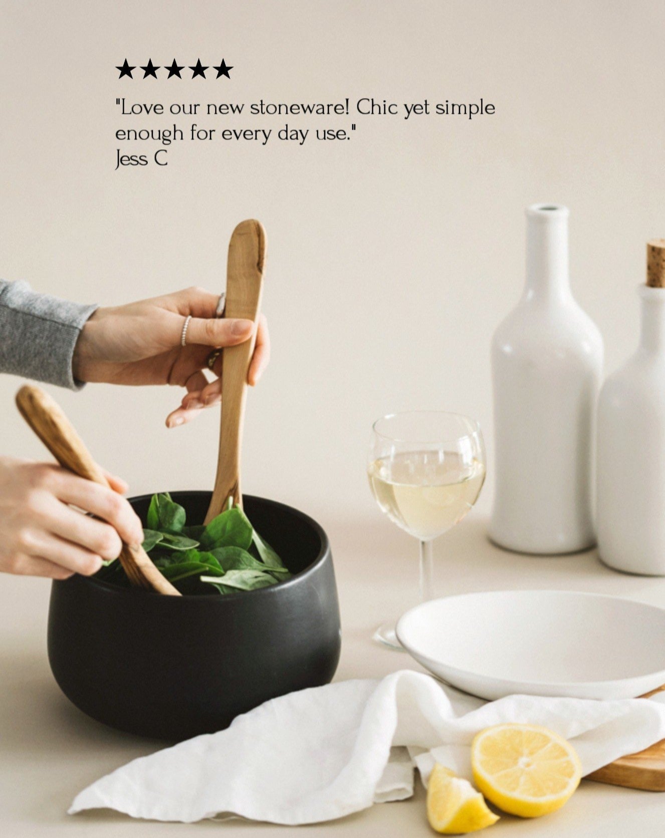 A person is serving salad from a black stoneware serving bowl on a dining table, with a white plate and a glass of white wine in the background.