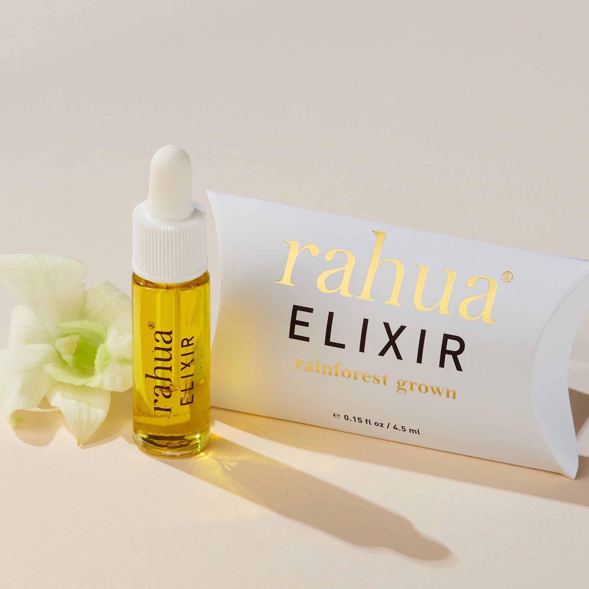 bottle of golden liquid in a glass bottle rahua elixir oil made of 100% rahua oil on a beige background with its pillow pack with gold rahua logo and elixir in brown|variant:015-oz