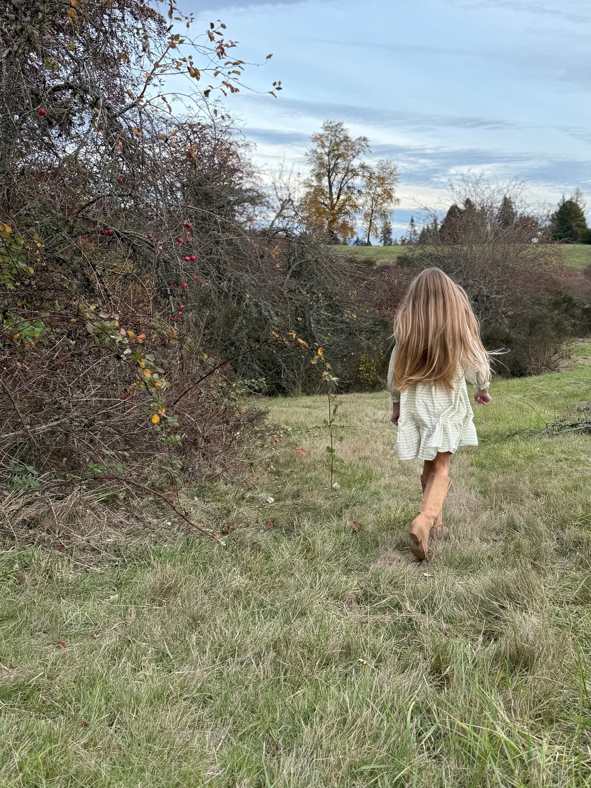 Person with long hair walking through a grassy field with trees and a blue sky in the background
