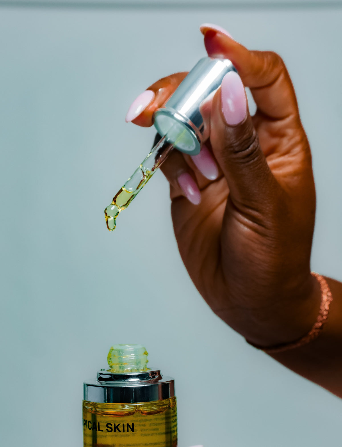 A close-up of a hand with pink manicured nails holding an open dropper bottle above another bottle, with a drop of golden-yellow serum dripping between the dropper and the container. The background is a solid light gray-blue.
