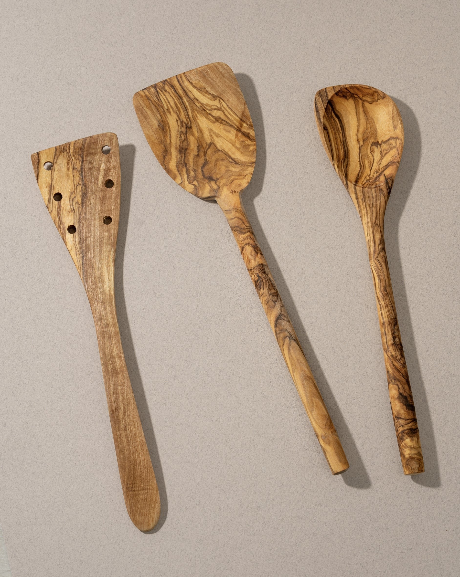 A set of three olive wood kitchen utensils, including a flipper, a pointed spoon, and a pancake spatula, laid out on a neutral surface.