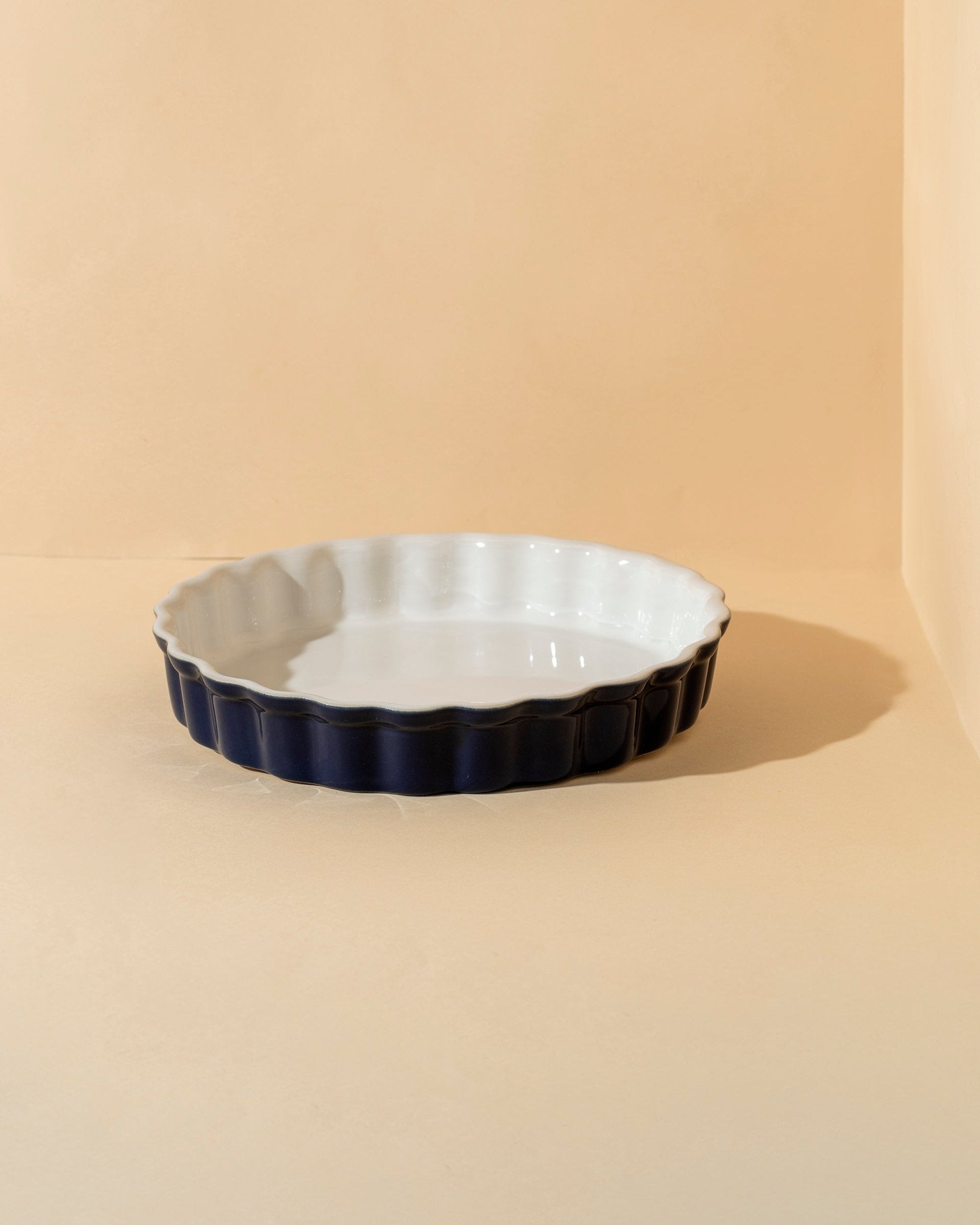 A white rounded stoneware pie dish with wavy side walls displayed on a neutral background, accompanied by a wooden cutting board with a pie on it, some garlic bulbs, and a small white bowl.