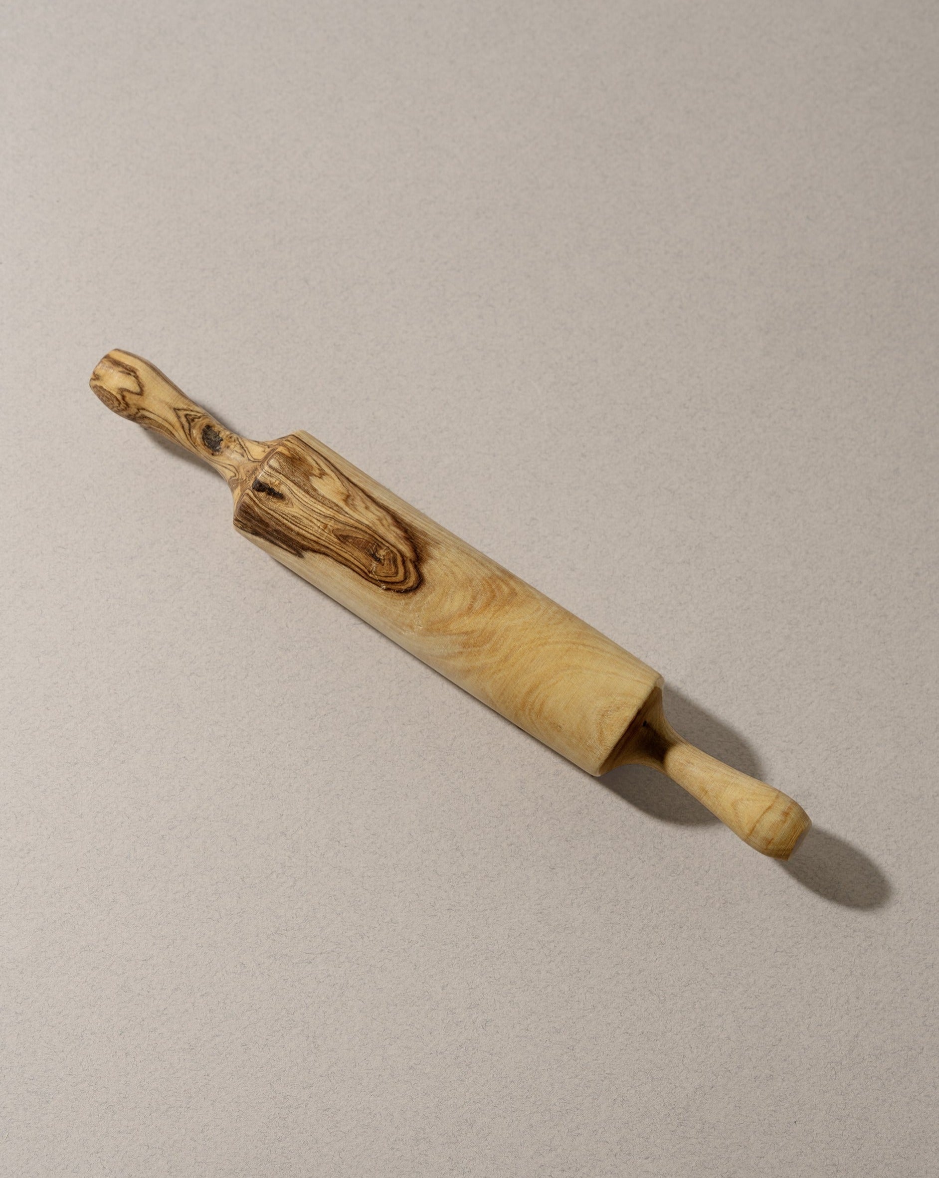 A single natural olive wood rolling pin displayed on an off-white background, highlighting its smooth finish, organic shape, and distinctive wood grain.