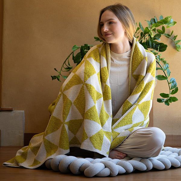 Woman wrapped in a green and white blanket with geometric patterns, sitting on a knotted floor cushion with plants in the background.