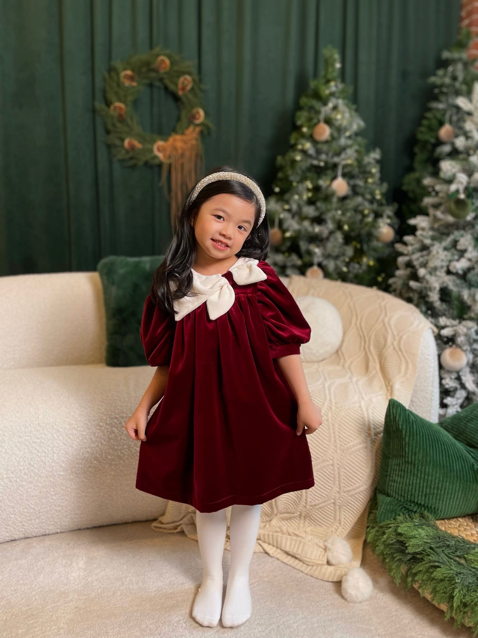 Young girl in a burgundy dress with white collar standing in a festive room with Christmas trees and decorations.