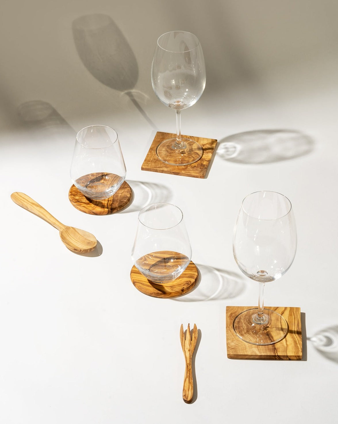 Two round and two square olive wood coasters, each holding a glass, arranged on a white background. A wooden spoon and fork are placed beside them, creating a cohesive natural dining setup.
