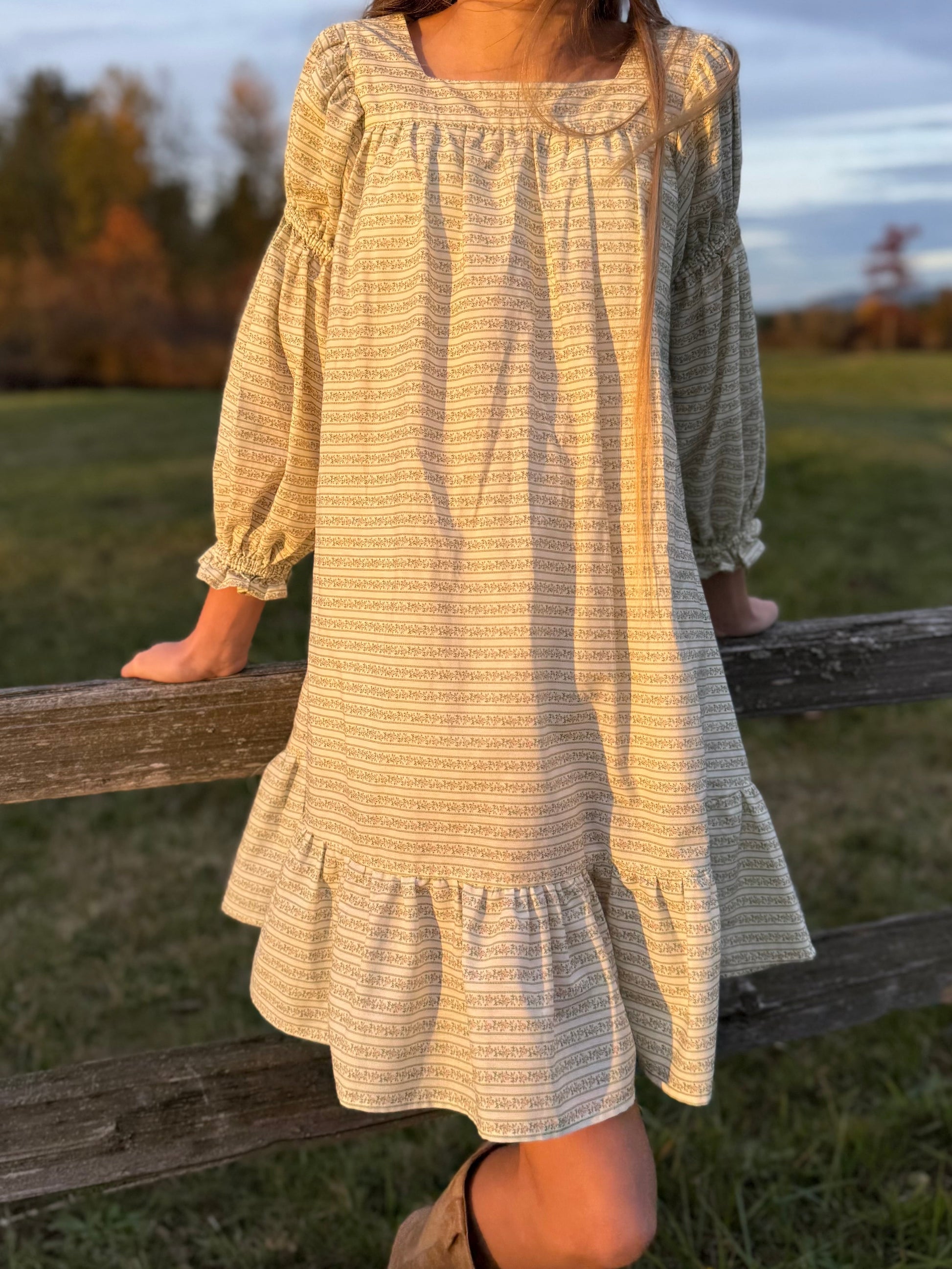 Person wearing a long, light-colored dress standing on a wooden fence in an outdoor setting.