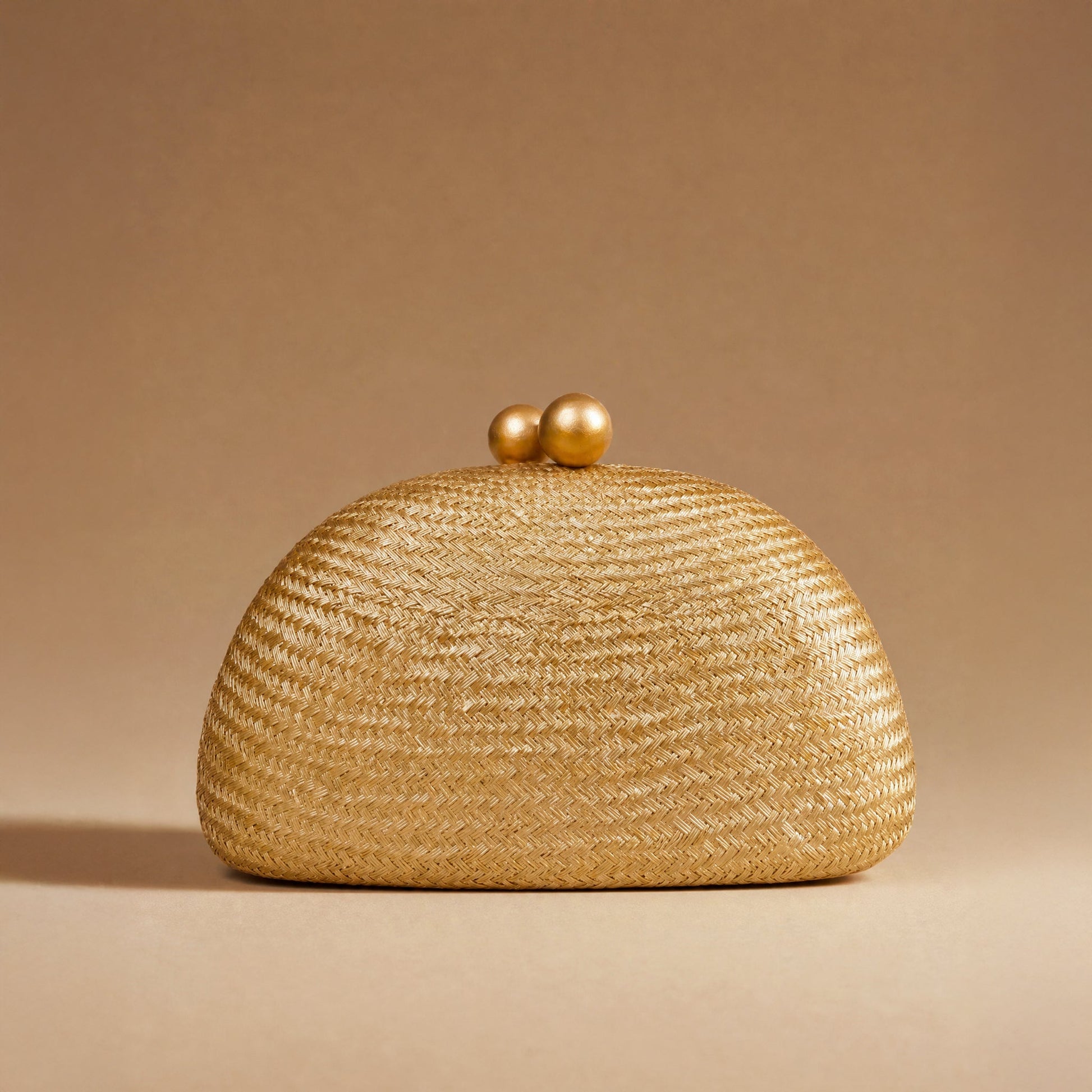A woven straw clutch purse in a warm golden-beige color, featuring a rounded dome shape and two small wooden ball clasp, set against a solid beige background.