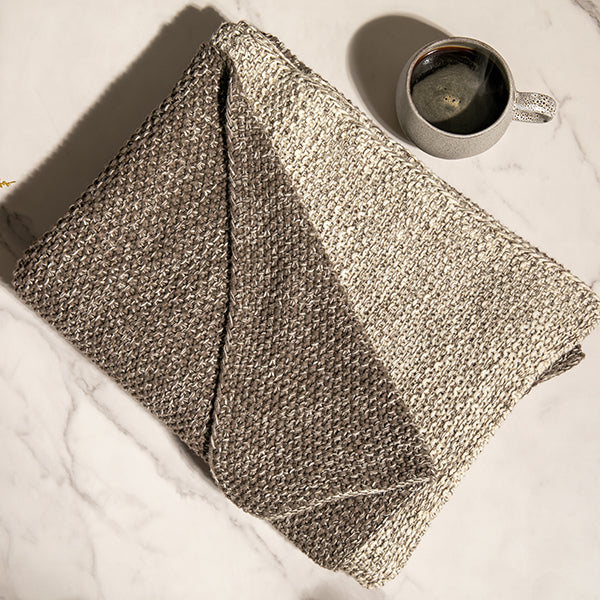 Folded reversible dark brown knitted throw cotton blanket on a white floor next to a cup of coffee.