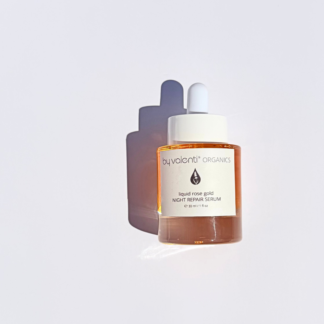 By Valenti glass bottle of vibrant orange/pink Liquid Rose Gold Night Repair Serum with a dropped on a violet table with bright midday sunlight