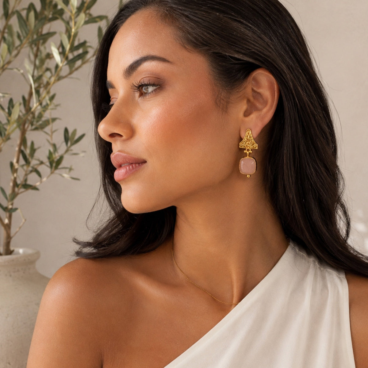 A woman in profile wearing a gold-plated statement earring with a rose quartz gemstone, against a soft beige background with a blurred olive plant.