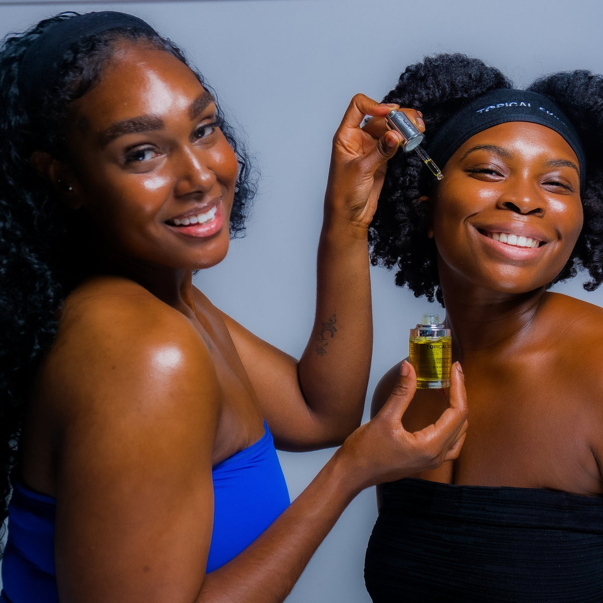Two Black women smiling in a studio setting, one holding a small vial of golden-yellow liquid and applying it to the other woman's face with a dropper. The woman on the left wears a bright blue strapless top, and the woman on the right wears a black strapless top and a black headband.