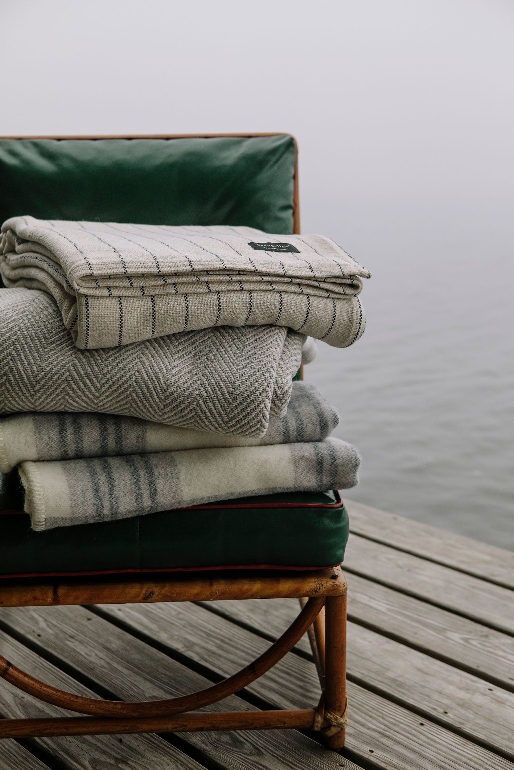 A luxurious stack of Evangeline Linens blankets, showcasing a timeless pinstripe pattern, rests gracefully on a wooden chair by the tranquil water. Crafted from 100% Cotton, these bed blankets are available in king, queen, and twin sizes. With their durability and washable nature, they are not only a symbol of luxury but also an heirloom to be cherished.