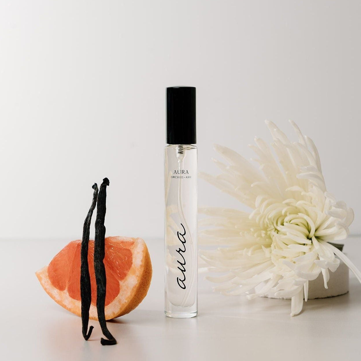 Perfume bottle with vanilla beans, grapefruit, and a white flower on a light background