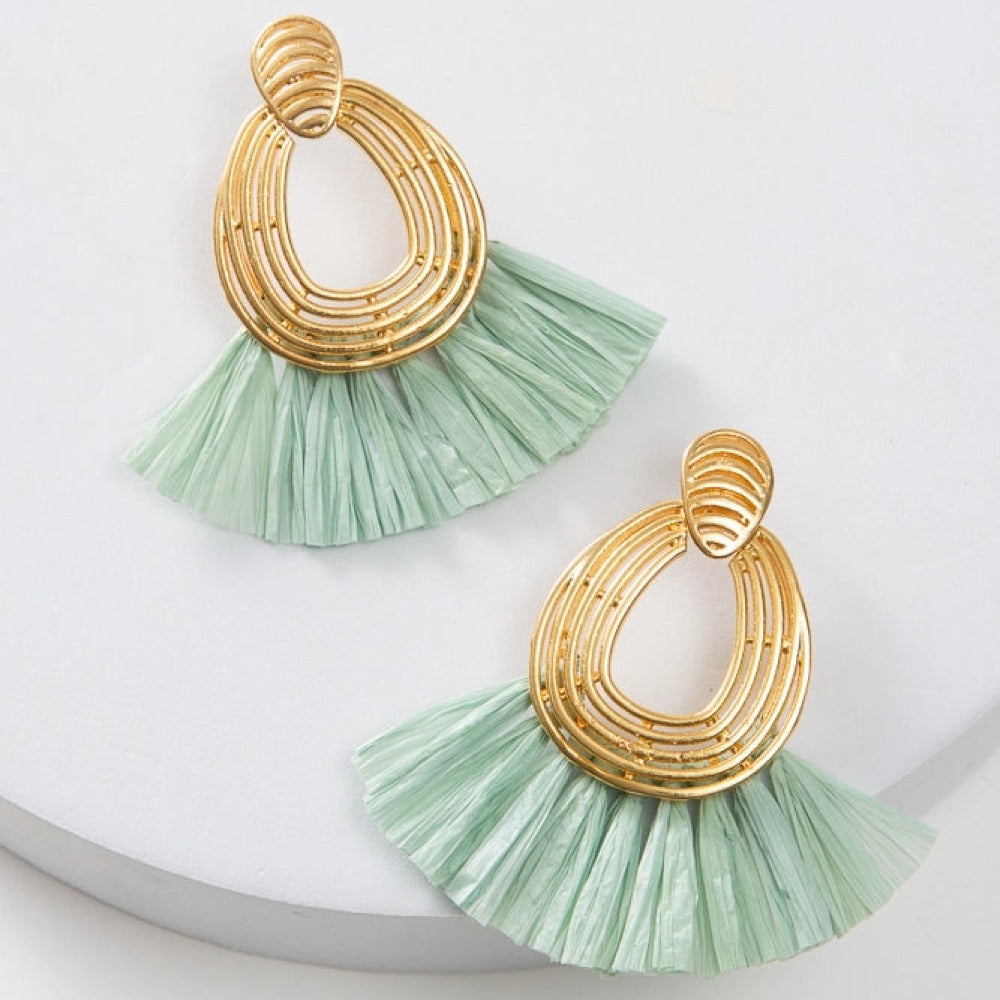 The Lux Raffia Earrings are post-style hoop earrings. The hoop is composed of thin rods of brass molded into an oval shape that is open in the center. Mint colored raffia fringe fans out from the bottom of the hoop.