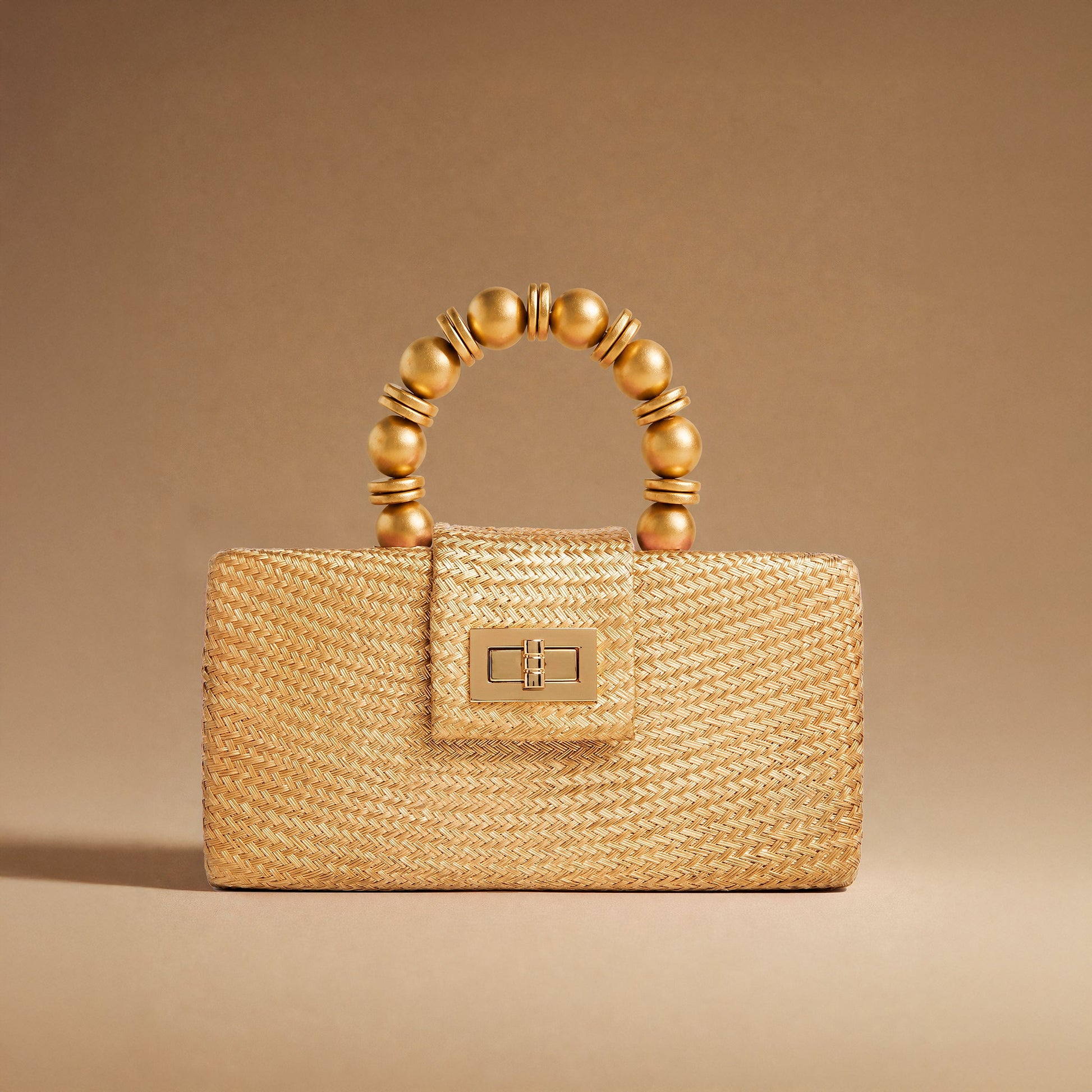 A structured woven straw handbag in a warm gold tone, featuring a curved top handle made of glossy gold-toned beads and a matching gold-toned clasp on the front flap, set against a solid warm brown background.