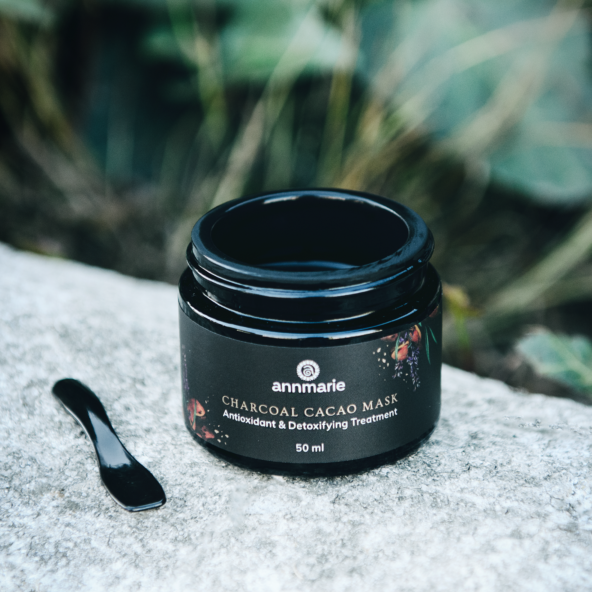 Black jar of annmarie Charcoal Cacao Mask on a textured surface with greenery in the background