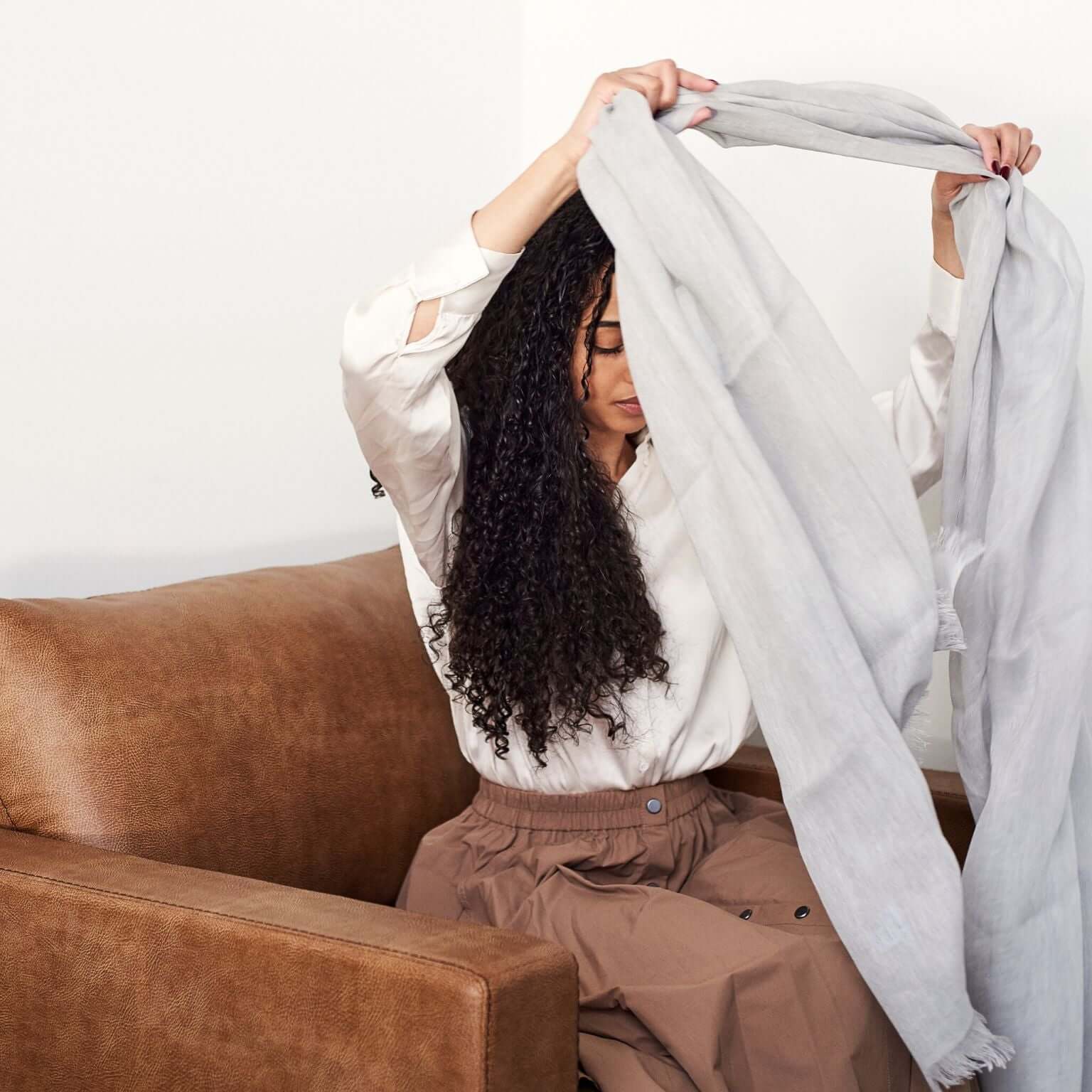 Woman styling Pebble Light Gray Hand Woven Soft Linen Scarf while sitting on a couch.
