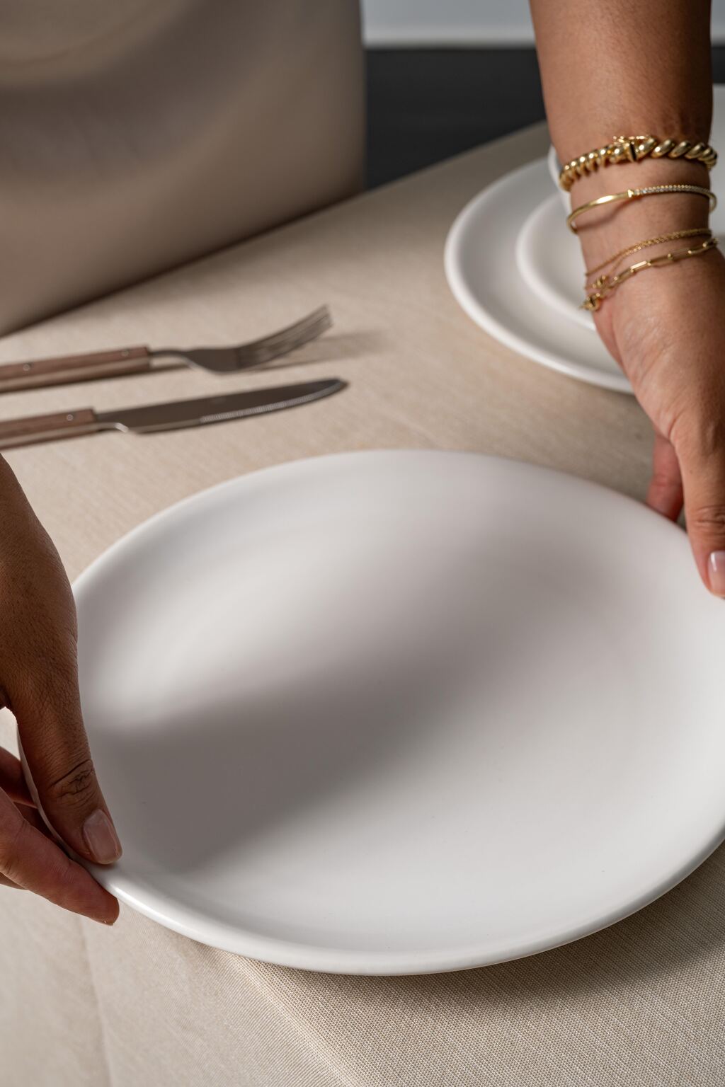 Bone-colored ceramic dinner plate from Hamkke Home being set on a linen tablecloth with wooden-handled flatware nearby.