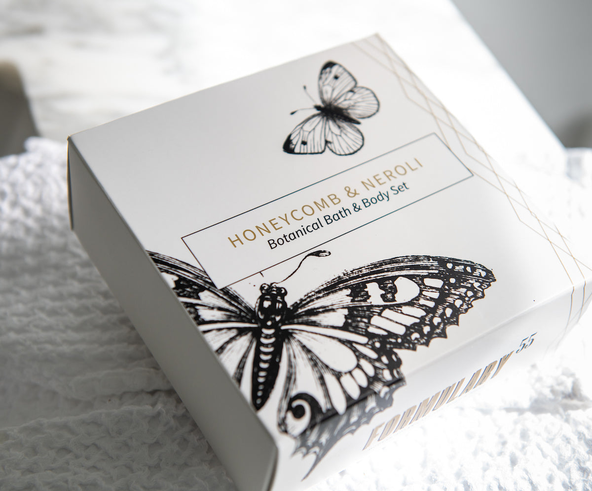 A white box with black illustrations of butterflies contains a "Honeycomb & Neroli Botanical Bath & Body Set." The box is resting on a textured white surface. The packaging design is elegant and minimalist.