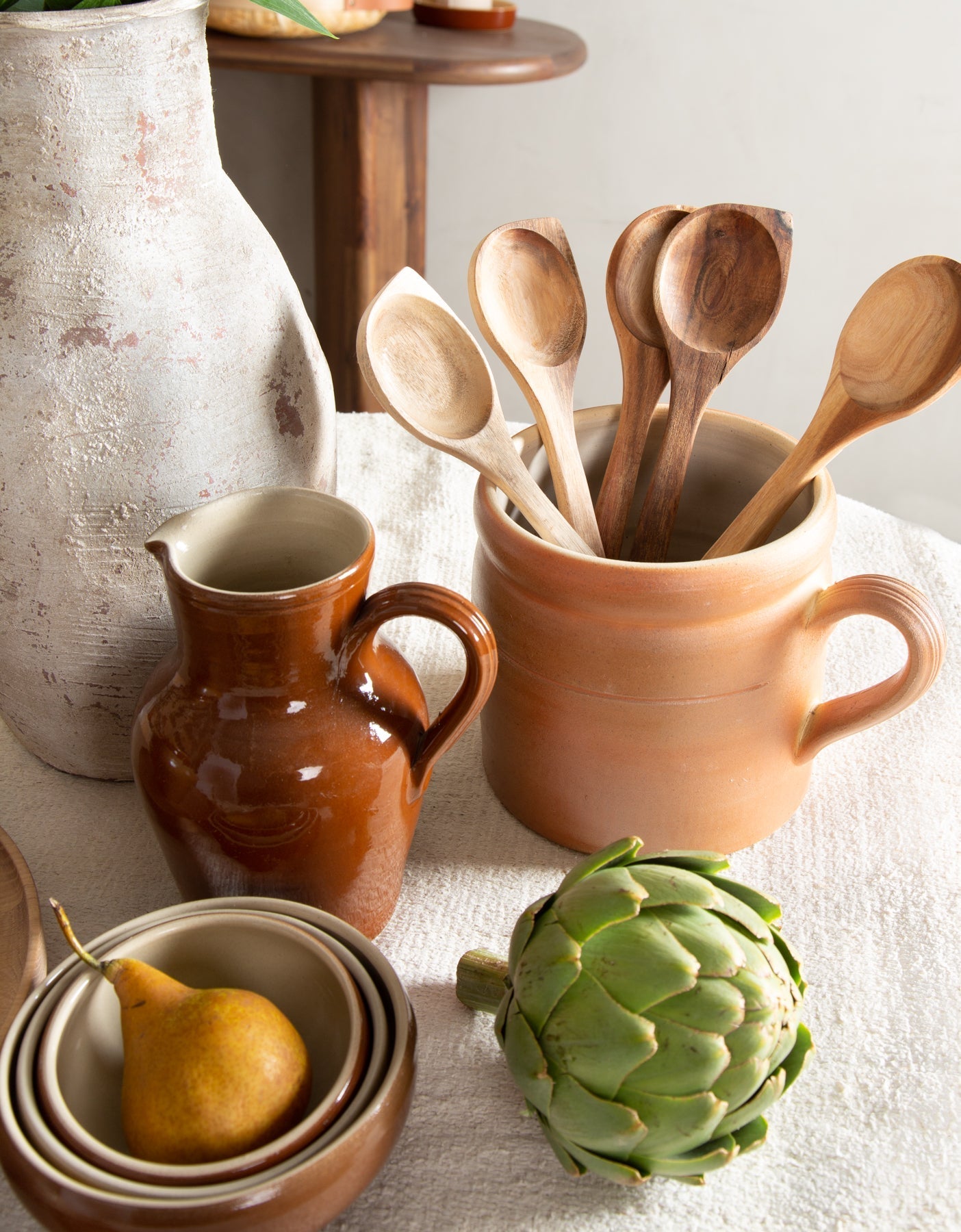 Set of wooden spoons in a ceramic container with ceramic jugs and a pear on a light surface.